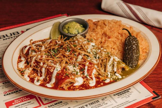 A plate of enchiladas topped with red and green sauce, crumbled cheese, and crispy tortilla strips. Sides of rice, guacamole, a pickled jalapeño, and a lime wedge accompany the dish. A menu and napkin are visible in the background.