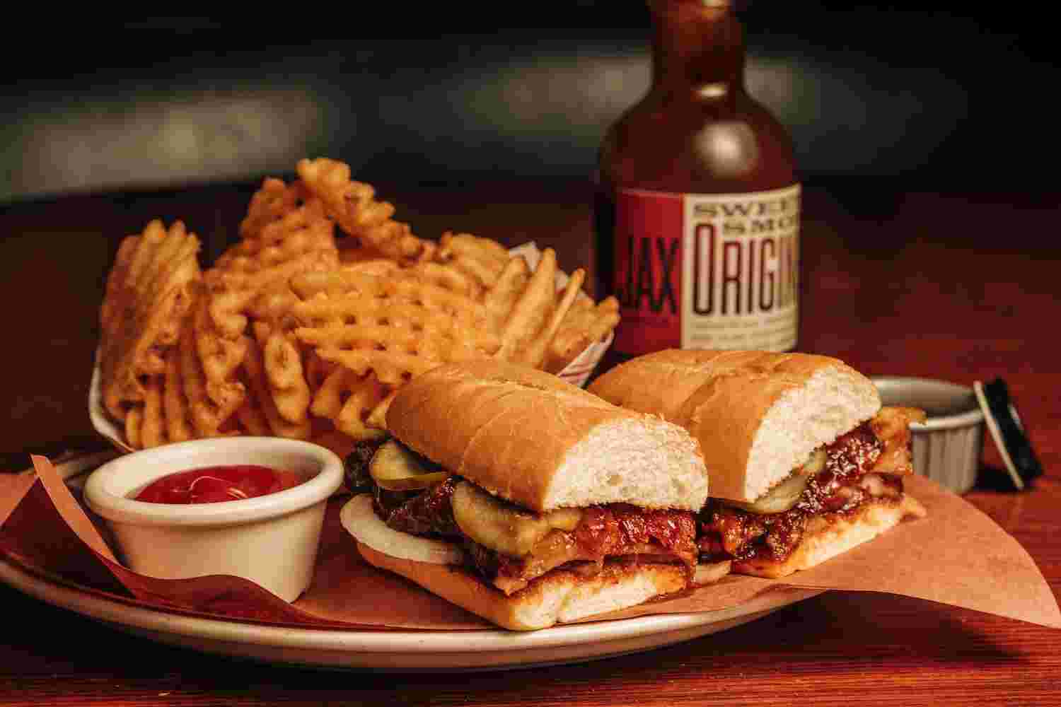 a bbq sandwich served with a side of ketchup, waffle fries, and a bottle of Jax's BBQ sauce