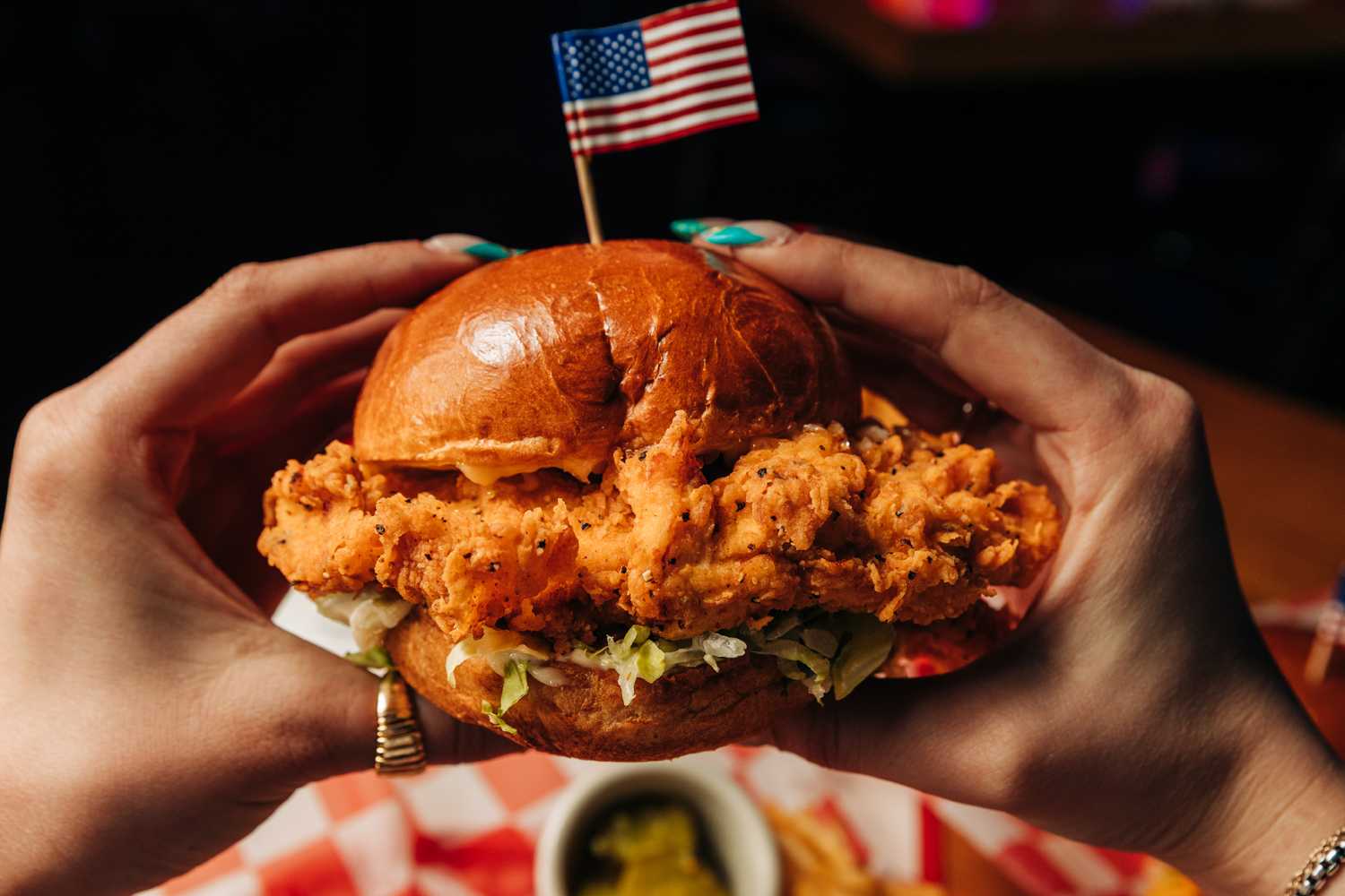 Two hands holding a Fried Chicken with lettuce sandwich on a bun with an American Flag toothpick in it. Red checkered paper is visible underneath.