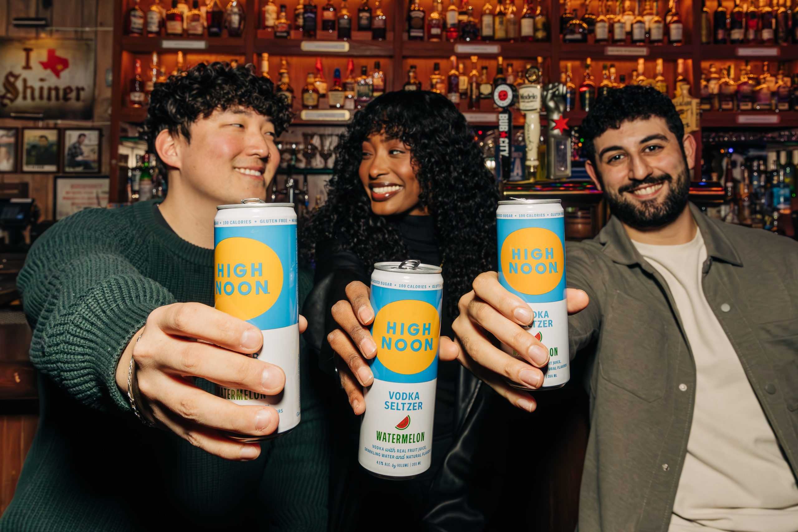 Three people holding out cans of high noon and smiling in a bar environment with a wall full of bottles behind them.