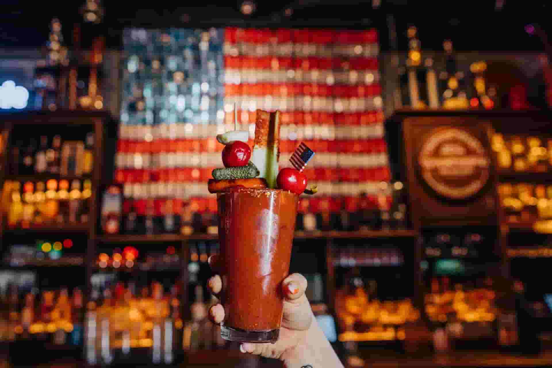Bloody Mary cocktail in a glass being held up by a hand in front of Bub City's beer can American flag