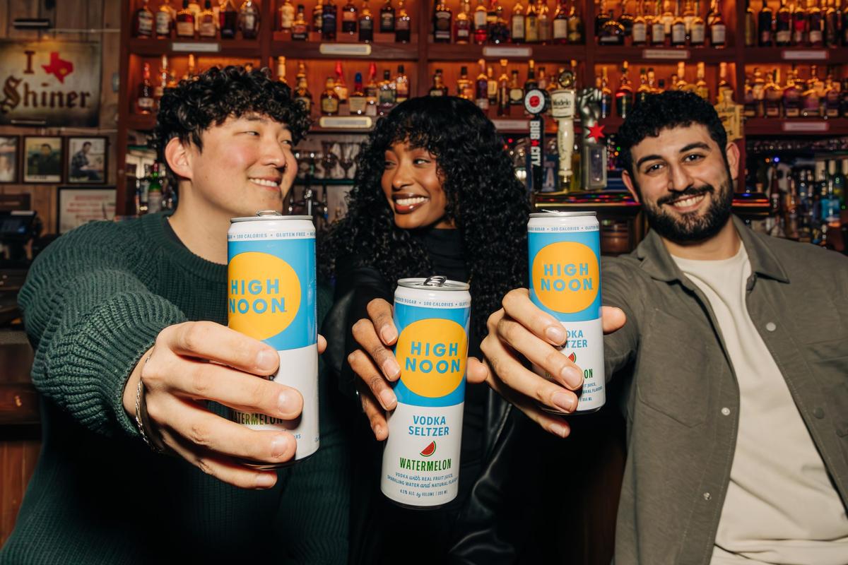 Three people holding out cans of high noon and smiling in a bar environment with a wall full of bottles behind them.